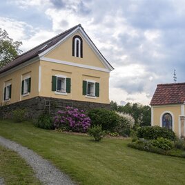 Ein gelbes Haus mit grünen Fenstern steht auf einem hügeligen Grundstück. Neben dem Haus befindet sich eine kleine Kapelle umgeben von bunten Blumen. | © Troadkammer | Fam. Stelzl