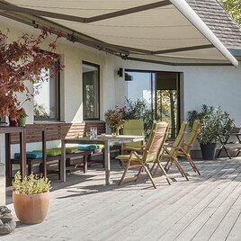 Eine moderne Terrasse mit Holzfußboden, Stühlen und einem Tisch. Im Hintergrund sieht man ein schönes Haus und grüne Pflanzen. | © Hertha Hurnaus
