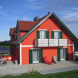 Ein schönes rotes Haus mit grünen Fensterläden und einem Balkon. Der Garten ist gepflegt und hat Terrassenmöbel. | © Ferienhaus Goriup