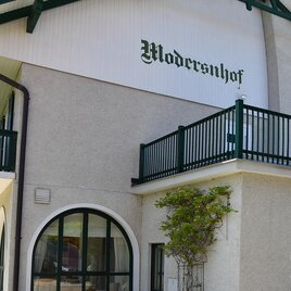 Ein modernes Gebäude mit einem Balkon und dem Schriftzug "Modernshof". Die Fassade ist hell und freundlich mit Pflanzen in der Nähe. | © Modersnhof