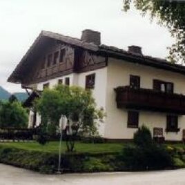 Ein traditionelles Haus mit einem Holzbalcony und einem gepflegten Garten. Im Hintergrund sind Berge und eine bewölkte Himmel zu sehen.