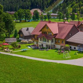 A large, traditional building with a red roof, surrounded by a green meadow. There are play areas and a beautiful view of the landscape.