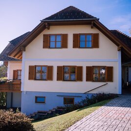 A beautiful house with brown shutters and a clear sky. The garden is well-maintained and the driveway is paved. | © Kurkommission Bad Blumau