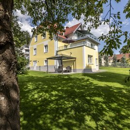 Gästehaus Landgraf in Üblbach mit gelber Fassade, Terrasse und grünem Garten mit Bäumen im Schatten. | © TV Region Graz-J