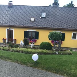 Ein gelbes Haus mit grünen Fensterläden und bunten Blumen in Töpfen. Der Garten ist mit Hecken und einem weißen Ball dekoriert. | © Gästezimmer Sabathi | Gertrude & Gottfried Sabathi