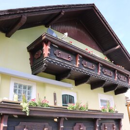 A traditional house with a wooden balcony adorned with flowers. The facade is painted in soft shades of yellow.