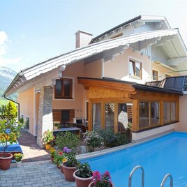 A modern house with a beautiful swimming pool and flower pots. In the background, the mountains and a blue sky can be seen.