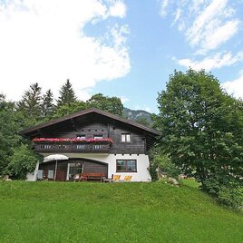 A charming wooden house with a balcony decorated with flowers. Surrounded by green grass and trees under a clear sky.
