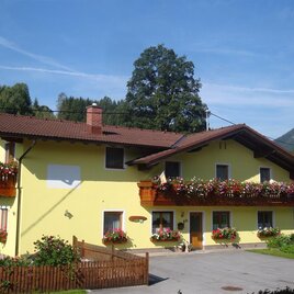 A cozy yellow house with blooming balcony flowers. In the background, green mountains and a blue sky can be seen.