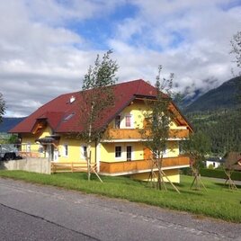 A yellow house with a red roof stands at the roadside. In the background, green mountains and trees are visible.