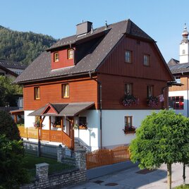 A charming wooden house with a red paint in a picturesque setting. In the background, green hills and a small church can be seen.
