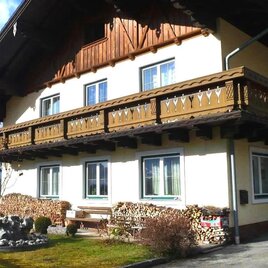 Ein traditionelles alpenländisches Haus mit einem Holzbalkon. Der Garten ist gepflegt und es gibt Holzstapel neben dem Gebäude.