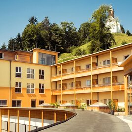 Ein modernes Hotelgebäude mit orangefarbenen Fassaden. Im Hintergrund ist ein grüner Hügel mit einem Leuchtturm zu sehen. | © Lupi Spuma