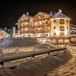 Ein malerisches Hotel in einer verschneiten Landschaft, beleuchtet von warmem Licht in der Dunkelheit. Der Weg zum Hotel ist von Schnee und einem Holzzaun umgeben.