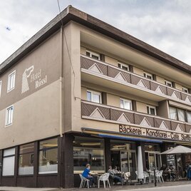 Ein großes Gebäude mit einem Café und einer Bäckerei im Erdgeschoss. Die Fassade ist einfach und modern, umgeben von Sitzplätzen im Freien.