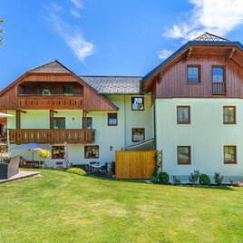 Ein schönes Haus mit Holzfassade und Garten. Im Hintergrund sind grüne Hügel und blauer Himmel zu sehen.
