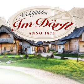 Ein malerisches Dorf mit traditionellen Holzhäusern und einem gepflegten Garten. Der Schriftzug „Wohlfühlen Im Dörfl Anno 1873“ verleiht dem Bild einen historischen Charme.