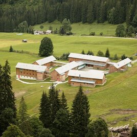 Eine ruhige Landlandschaft mit mehreren Holzbauten und grünen Wiesen. Umgeben von dichten Waldflächen und sanften Hügeln. | © JUFA Hotel Eisenerz