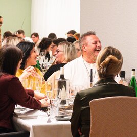 A lively restaurant atmosphere with several guests sitting at a table and laughing. There are glasses and bottles on the table, indicating a sociable meal. | © Kunsthotel Kirchenwirt