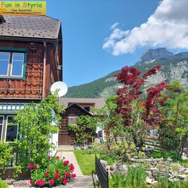 A picturesque house surrounded by colorful flowers and green trees. In the background, majestic mountains and a blue sky can be seen.