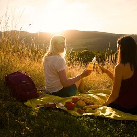 Two friends are enjoying a picnic on a meadow at sunset. The evening sky glows warmly, and the landscape is picturesque. | © Oststeiermark Tourismus