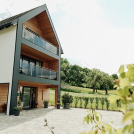 Ein modernes Haus mit großen Fenstern und einem Balkon. Die Umgebung ist grün und lädt zum Entspannen ein.