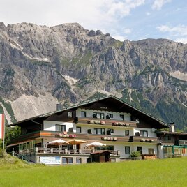 A traditional Alpine house with a beautiful porch. In the background, majestic mountains rise under a clear sky. | © Herold