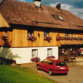 A traditional wooden house with flower boxes and a red car in front. The sky is partially cloudy.