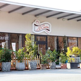 Ein Restaurant mit einem bayerischen Flair und einer Bäckerei im Vordergrund. Die Außenansicht ist mit Pflanzen dekoriert. | © La Cucina