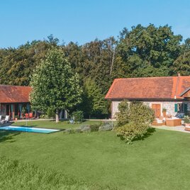 Ein modernes Haus mit rotem Dach und großem Garten. Im Garten befindet sich ein Pool und hohe Bäume umgeben die Anlage.