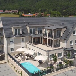 A modern building with several floors and balconies. In the foreground, there is a swimming pool and cozy seating areas. | © Vulkan TV