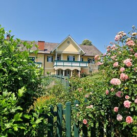 Ein charmantes Haus mit einem bunten Garten und vielen Blumen. Im Hintergrund strahlt ein klarer blauer Himmel. | © KK