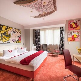 Ein modernes Hotelzimmer mit einem Doppelbett und bunten Kunstwerken an den Wänden. Der Boden ist mit orangefarbenem Teppich ausgelegt und es gibt einen kleinen Tisch mit einer Vase voller Blumen.