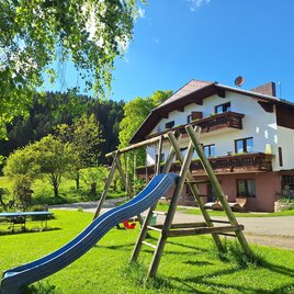 An idyllic house surrounded by green meadows and trees. In the foreground, there is a slide on a playground.