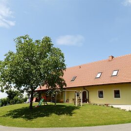 Ein schönes Haus mit rotem Dach und großer Wiese. Ein Baum steht neben dem Haus und die Umgebung ist grün und einladend. | © Weinbauhof Bernhard | Fam.Bernhard