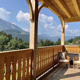 Ein Balkon mit Holzgeländer und zwei Stühlen, umgeben von grünen Bäumen und Bergen. Der Himmel ist blau mit wenigen Wolken. | © Julie Albers
