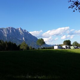 Eine malerische Landschaft mit majestätischen Bergen und einem klaren blauen Himmel. Im Vordergrund erstreckt sich eine grüne Wiese mit vereinzelten Bäumen.