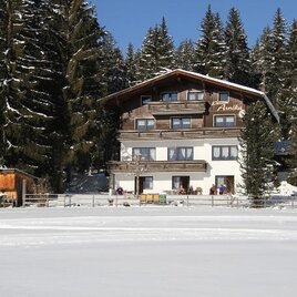 Ein gemütliches Haus im Schnee mit umgebenden Nadelbäumen. Der klare blaue Himmel sorgt für eine malerische Winterlandschaft.