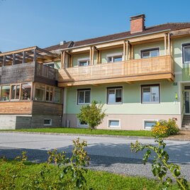 Ein modernes Gebäude mit einem Holzbalustrade und einem Glasbalkon. Die Fassade ist in sanften Farben gehalten und von grünen Pflanzen umgeben. | © Pension Cäcilia