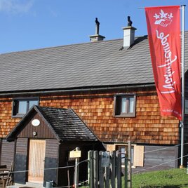 Ein rustikales Holzgebäude mit einem grauen Dach und einem roten Banner. Es befindet sich in einer ländlichen Umgebung, umgeben von einer Wiese. | © Petzhütte, www.naturfreunde-huetten.at/steiermark/pelzhuette/