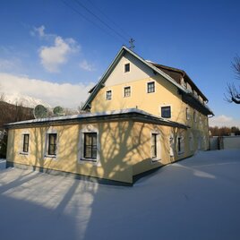 Ein gelbes Gebäude steht in einer verschneiten Landschaft. Der Himmel ist klar und blau, und die Schatten der Bäume sind auf dem Schnee zu sehen.
