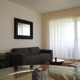 Ein modernes Wohnzimmer mit einem grauen Sofa und einem großen Spiegel an der Wand. Große Fenster mit weißen Vorhängen lassen viel Licht herein. | © Relax Residence Südsteiermark | Reinhard Neussner