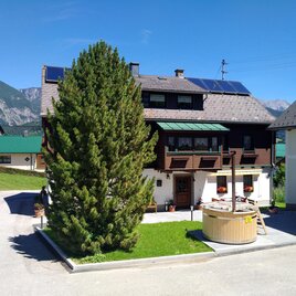 A cozy house with a large tree in the foreground. In the background, mountains and a clear blue sky can be seen.