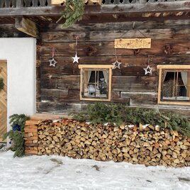 Ein rustikales Holzhaus mit einer dekorativen Eingangstür. Vor dem Haus liegt ein Stapel Holz und der Boden ist mit Schnee bedeckt.