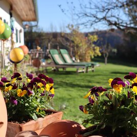 Ein sonniger Garten mit bunten Blumen in Töpfen. Im Hintergrund stehen Liegestühle und eine entspannte Atmosphäre ist sichtbar. | © Sonnenhaus Grandl