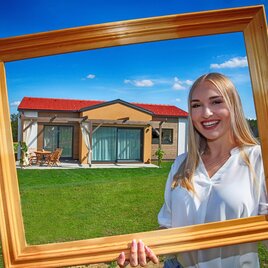 Eine lächelnde Frau hält einen goldenen Rahmen, der ein modernes Haus zeigt. Im Hintergrund sind eine grüne Wiese und ein klarer Himmel zu sehen. | © Sepp Pail
