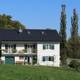 Ein modernes, zweistöckiges Haus mit einem schwarzen Dach und grünen Fensterläden. Es liegt auf einem sanften Hügel, umgeben von Bäumen und einem gepflegten Garten.
