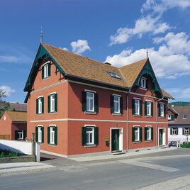 Ein hübsches rotes Haus mit grünen Fensterrahmen und einem schrägen Dach. Die Umgebung zeigt eine ruhige Straße und andere Gebäude. | © Villa Franziska/Helmut Schweighofer