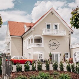 Eine elegante Villa mit einem roten Oldtimer vor der Haustür. Der gepflegte Garten und die stilvollen Bäume verleihen dem Anwesen einladenden Charme. | © Villa Thermale