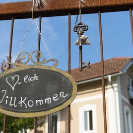 Ein herzlicher Willkommensschriftzug hängt an einem Zaun. Im Hintergrund ist ein Gebäude mit einem roten Dach zu sehen. | © Villa zur Schmied'n | Petra Zirngast
